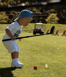 Child holding a black adjustable golf driver club. Child about to hit an orange golf ball on the green grass of a driving range. Yellow toddler tee on the ground near the golf ball. Blurred golf cart in the background. Mini Hoshi branded golf club.