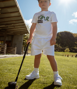 Child in a white outfit with a Mini Hoshi branded t-shirt, holding a black adjustable golf driver club. Standing on a grassy area with trees, hills and a driving range building in the background. Mini Hoshi branded golf club.