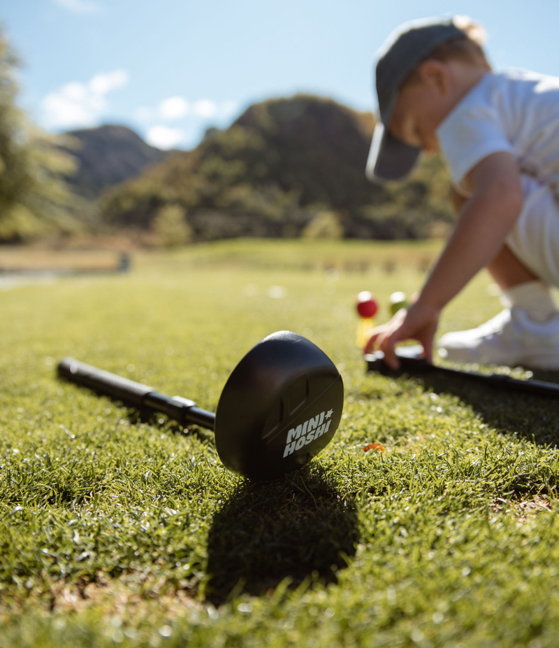 Black adjustable toddler golf driver club, lying on the green grass with the head close up and visibly branded with a white Mini Hoshi logo. Child blurred in the background about to pick up a golf putter. Two golf balls (red and green) visible in the background. Set on a driving range with blurred mountain in the background. Mini Hoshi branded golf club.