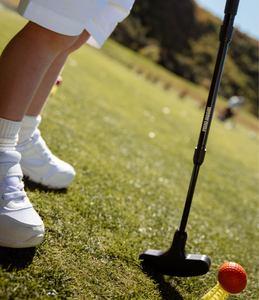 Young child holding a golf putter, with yellow plastic tee holding a bright orange golf ball. Image on an angle, close up and cropped with a blurred background.