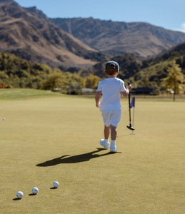 Child on a golf course with mountains in the background walking towards a golf green holding an adjustable putter. Golf balls in the foreground resting on the green. 