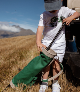 Child holding a green leather and canvas golf bag in a grassy field with mountains in the background. Child is pulling  a putter golf club out of the top of the golf bag. 