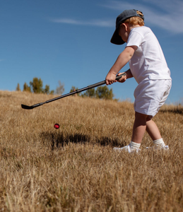 Child swinging an adjustable golf wedge at a bright red golf ball in in a field, with a clear sky blue sky. Mini Hoshi branded golf club. 