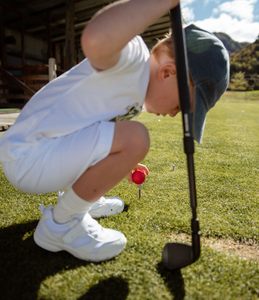 Child playing golf on the grass of a driving range, with a barn in the background. Child is crouching down to place a bright red golf ball on a tee and clutching an adjustable golf wedge club. Mini Hoshi branded golf club. 
