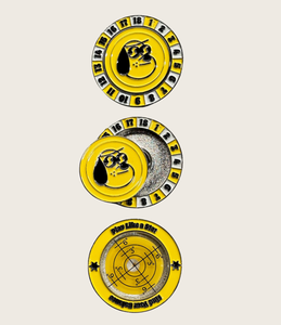 Bright yellow, round ball marker with numbers one to eighteen around the outside similar to a poker chip. The inside has a removable metal chip with an image of a black dog. The reverse image of the ball market shows a level and the words 'play like a star' and 'find your balance'. Mini Hoshi branded golf ball marker for toddlers. 