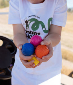 Child holding colourful Blue, pink, orange, yellow and blue golf balls with a white t-shirt featuring a character hitting a golf ball and the words 'I in the background.lay golf now' on the front. Mini Hoshi branded.