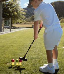 Child playing golf, holding an adjustable putter, on a sunny day with trees and mountains in the background. Child is about to hit one of three golf balls set up in a row on yellow toddler starter tee's. The colourful balls are red, green and red. Child s dressed in white. Mini Hoshi branded golf club.