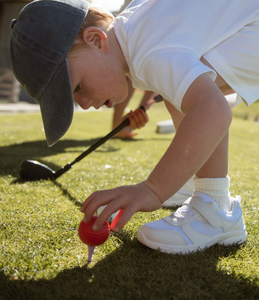 Child leaning down to the ground to carefully place a bright red gold ball onto a purple bamboo tee which is held in the ground. Child clutches an adjustable golf wedge club in his right hand. Mini Hoshi branded golf club.