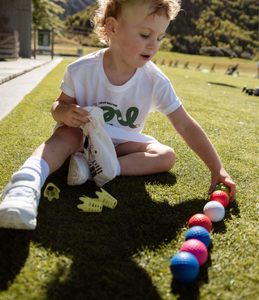 Child dressed in white sits on the grass at a driving range and is lining up colourful golf balls whilst holding a Mini Hoshi branded fabric bag. 