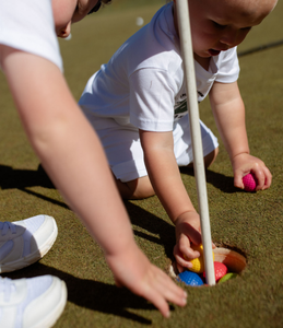Two children on the green at a golf course, reaching down to put colourful golf ball in the hole. 