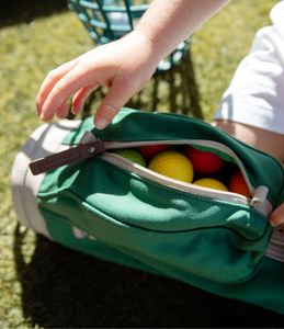 Green toddler golf bag with colourful balls inside the side pocket, zip is open and a small child's hand n reaching down to pull the zipper tag which is branded in the leather Mini Hoshi. Child is kneeling on green grass with a blurred golf basket int he background.