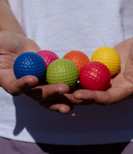 An adult with a white t-shirt is holding six balls in this hands. Close up image of a blue, pink, green, orange, red and yellow golf ball set. 