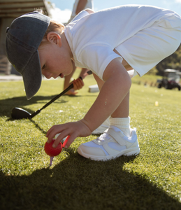 Child playing golf on a grassy driving range ground. Child is placing a bright red golf ball carefully on a purple golf tee. Child is holding a wedge club in their right hand. Blurred golf cart in the background. Mini Hoshi branded golf club. 